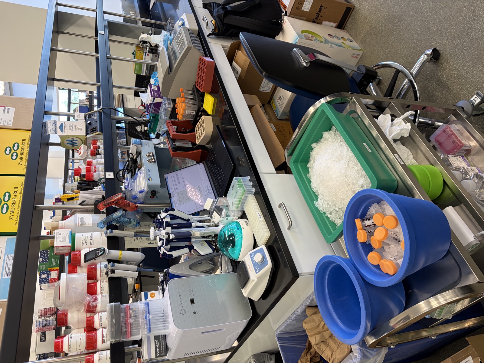 Crowded lab bench with equipment, reagents and a cart with ice buckets